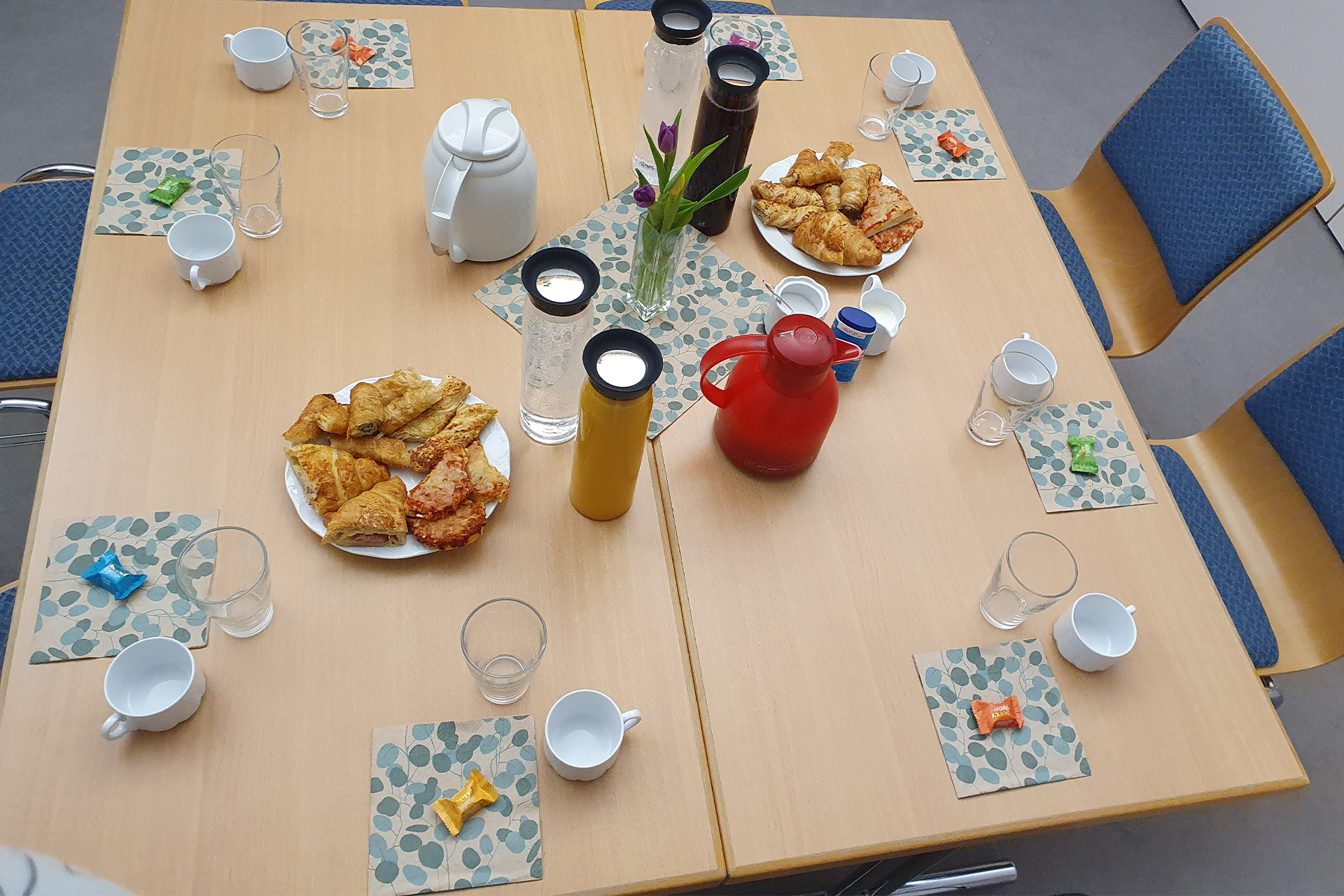 ein mit 8 Gedecken gedeckter Tisch. In der Mitte stehen Kaffee und Teekannen mit Gebaeck
