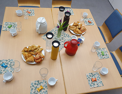 ein mit 8 Gedecken gedeckter Tisch. In der Mitte stehen Kaffee und Teekannen mit Gebaeck