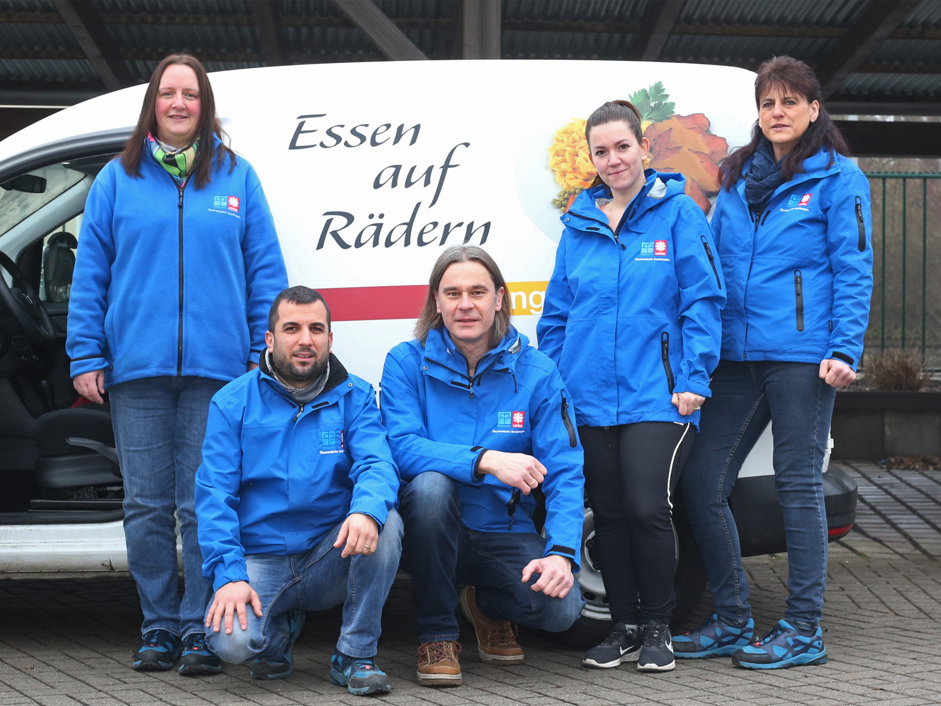 Fuenf Menschen stehen oder kauern vor einem Lieferauto mit der Aufschrift: 'Essen auf Raedern'