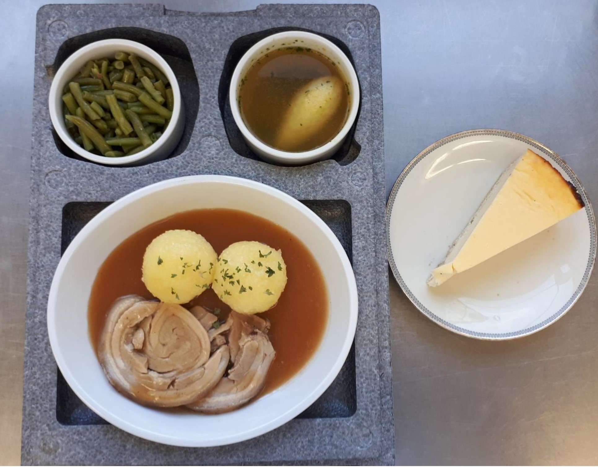 Es ist ein Essenscontainer mit einer Kompletten Mahlzeit zu erkennen. Diese besteht aus einer Hauptmahlzeit, Nachtisch und Suppe. Daneben steht ein Teller mit einem Stueck Kuchen