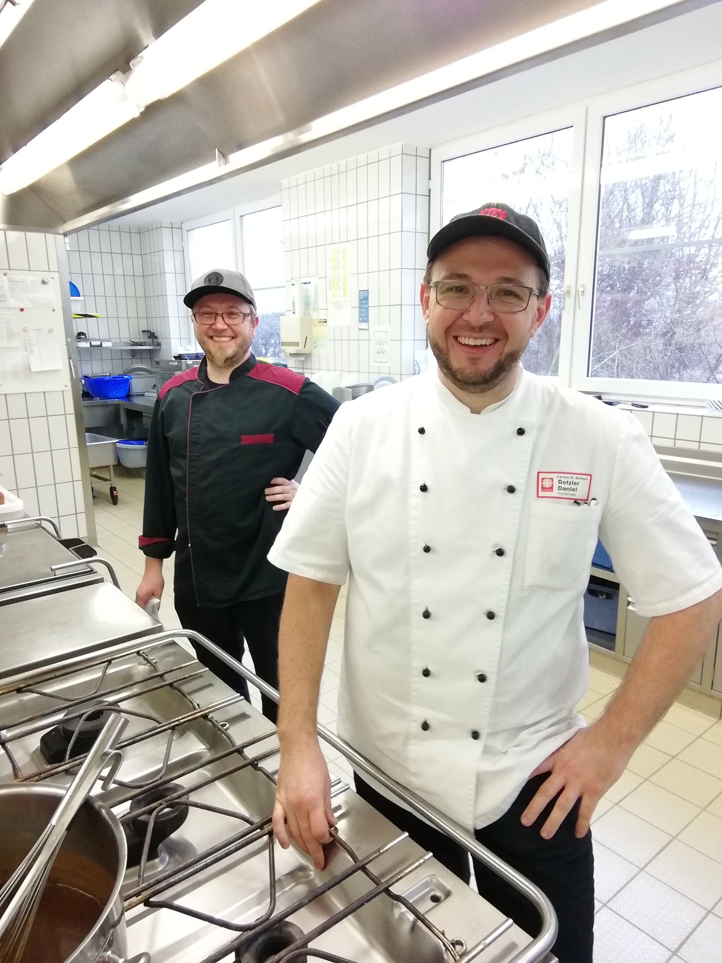 Zwei Personen stehen in einer Grosskueche vor einem Kueckengeraet und lachen in die Kamera