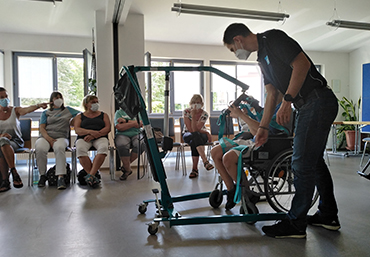 Es sind mehrere Personen in einer Stuhlreihe zu sehen. In der Mitte des Bildes ist eine Person, welche ein Pfleghilfsmittel erklaert und vorfuehrt.