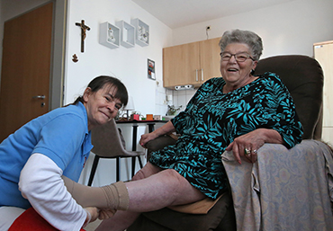 Eine aeltere Dame sitzt im Sessel. Vor ihr kniet eine Pflegeperson und zieht ihr die Struempfe an.