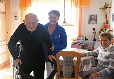 Ein aelterer Herr bekommt von einer Pflegeperson Unterstuetzung beim gehen mit dem Rollator. Eine Frau sitzt am dahinterstehenden Kuechentisch.