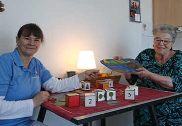 Zwei Personen, eine aeltere Dame und eine Pflegeperson sitezen an einem Tisch. Beide tauschen Utensilien zum Basteln aus.