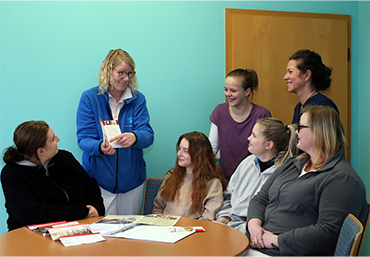Es sitzen und stehen mehrere Personen um einen Tisch. Auf dem Tisch liegen mehrere Informationsmaterialien. Eine Frau erklaert den Inhalt einer Broschuere