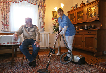 ein aelterer Herr sitzt am Tisch und eine Angestellte der Sozialstation saugt mit einem Staubsauger den Teppich im Wohnzimmer