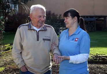 zwei Menschen stehen im Freihen. Die Frau des Pflegedienst hilft dem anderen Menschen beim gehen.