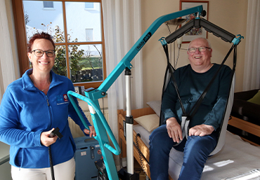 in einem Schlafzimmer wird ein aelterer Mensch mit einem Lifter von einer Pflegeperson aus dem Bett geholfen.