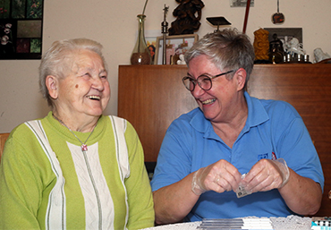 Zwei Menschen sitzen am Tische. Eine Pflegeperson bereitet Tabletten vor.