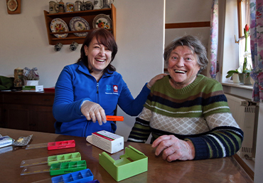 Zwei Menschen sitzen am Tische. Eine Pflegeperson bereitet Tabletten vor.