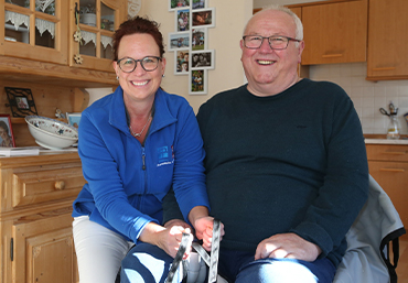 Zwei Personen sitzen in einer Kueche. Eine Pflegeperson haelt beispielhaft ein Hilfsmittel und erklaert dies der anderen Person